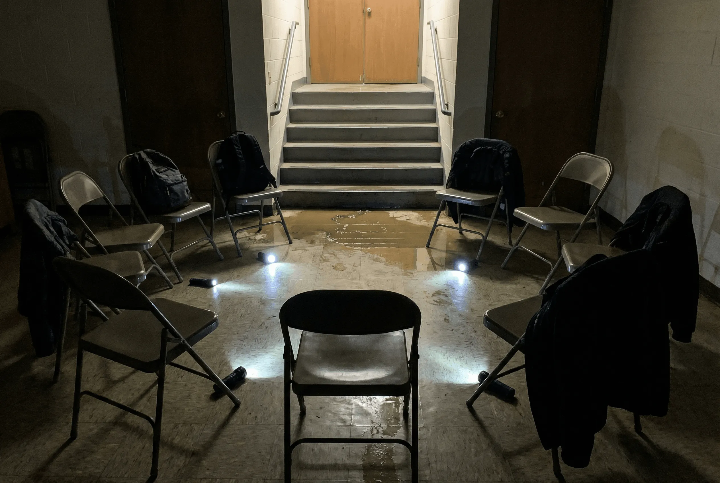 A dark church basement with folding chairs arranged in a circle, flashlight beams visible, water seeping under the door